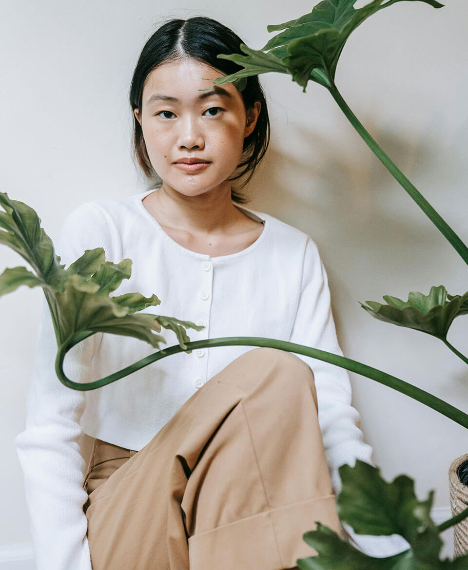 Femme assise en blanc et beige devant une plante verte, expression paisible.
