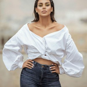 Femme debout, mains sur les hanches, chemise blanche à épaules dénudées.